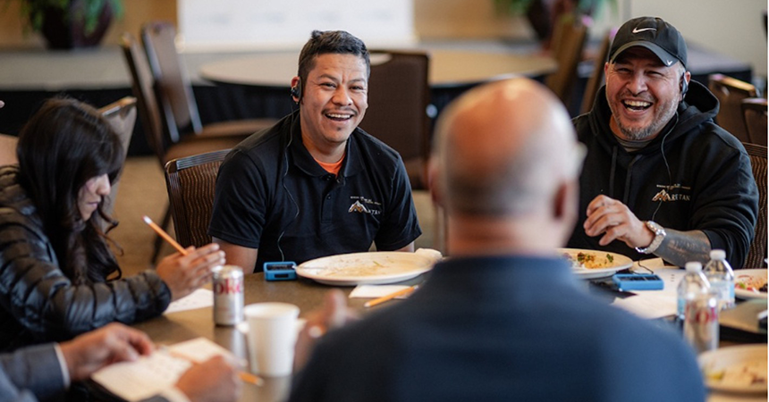 A group of casually dressed people sit around a rectangular table during an informal meeting or discussion. The table is cluttered with items including a can of Diet Coke, a white cup, a water bottle, papers, and plates with food remnants. One person writes on a notepad with a pencil. The background shows empty chairs and a softly blurred indoor setting.