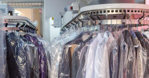 A circular dry cleaning conveyor system filled with garments on metal hangers, each covered in clear plastic bags. The clothes—mostly shirts and jackets—are neatly arranged and tagged with numbered labels for identification. The automated rack curves through the frame, showcasing an organized industrial setup typical of professional garment care facilities.
