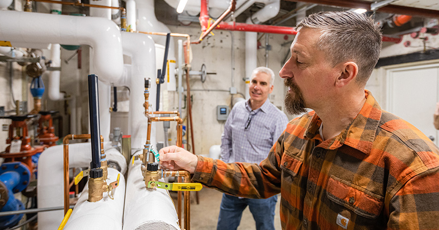 Two people in a mechanical room inspect HVAC and plumbing systems. One adjusts a valve on an insulated pipe while the other observes. The room contains exposed pipes, gauges, and industrial fixtures, suggesting maintenance or inspection work.