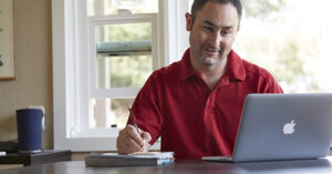 A person in a red polo shirt sits at a desk, smiling while working on a silver MacBook laptop. They hold a pencil and write in a notebook. A blue tumbler sits nearby, and sunlight streams through a window in the background, creating a bright, focused workspace.