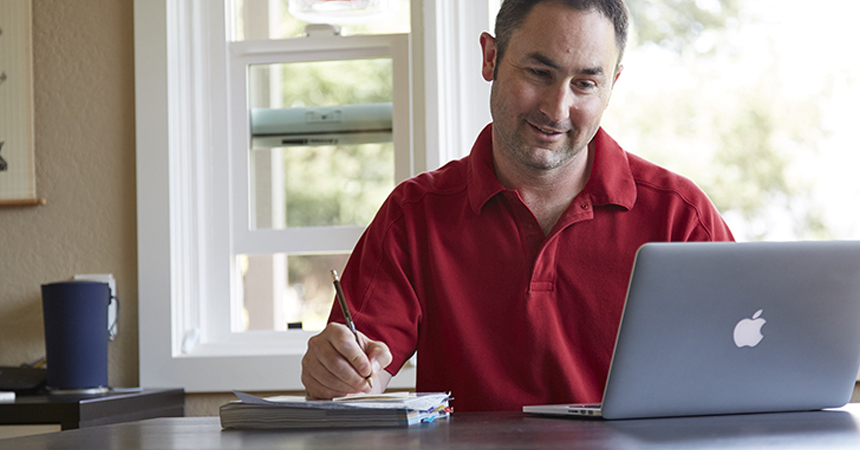 A person in a red polo shirt sits at a desk, smiling while working on a silver MacBook laptop. They hold a pencil and write in a notebook. A blue tumbler sits nearby, and sunlight streams through a window in the background, creating a bright, focused workspace.