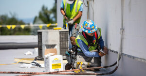 Two construction workers on a rooftop perform maintenance near a white wall. The foreground worker kneels, using a tool on a conduit, wearing a blue hard hat with stickers, blue shirt, safety vest, and harness. The background worker stands holding a tool, also in safety gear. Nearby are tools, cables, and a box labeled “VERSICO Roofing Systems – 850-SS White Cut-Off Mastic.” The scene highlights industrial roofing work and safety practices
