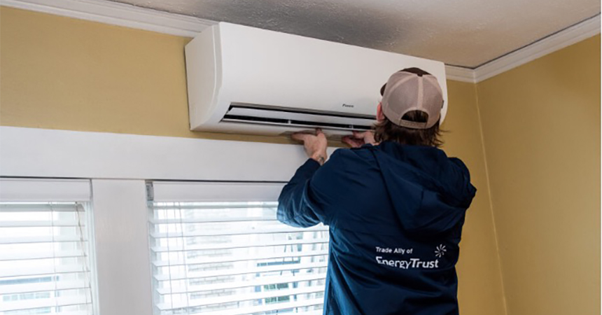 A person in a blue jacket and beige baseball cap services a wall-mounted air conditioning unit above a window with white blinds in a yellow-walled room. They are inspecting or adjusting the underside of the unit, highlighting a typical HVAC maintenance task.