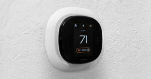 A sleek, modern ecobee smart thermostat mounted on a textured white wall, displaying a temperature of 71°F. The black front screen shows icons for Wi-Fi, lock, and flame, with a status message reading "1 Holding" below the temperature.