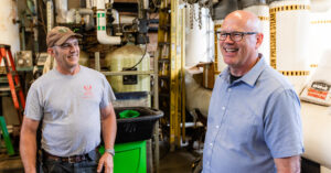 Two individuals stand in a mechanical room filled with industrial pipes and equipment. One wears a gray "University of Utah Facilities Management" t-shirt, the other a light blue button-up shirt and glasses. Both smile amid labeled pipes, tools, and a green trash bin, suggesting a friendly moment in a building maintenance setting.